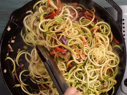 Zucchini noodles being stirred into skillet with cooked vegetables