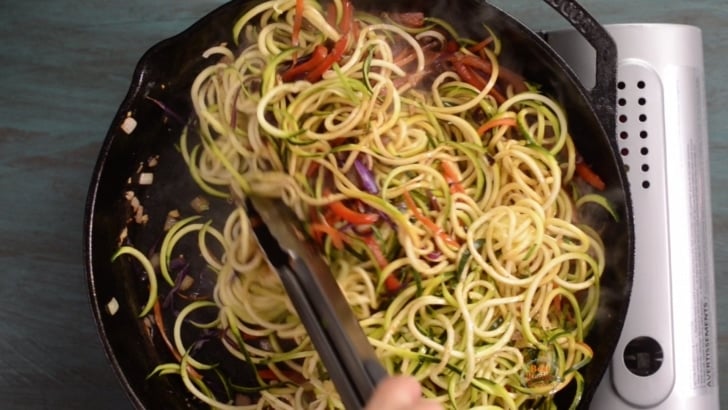 Zucchini noodles being stirred into skillet with cooked vegetables